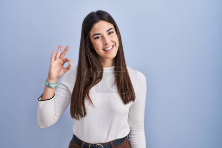 Foto de Young brunette woman standing over blue background smiling positive doing ok sign with hand and fingers. successful expression. - Imagen libre de derechos