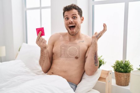 Photo for Young hispanic man holding condom in the bed celebrating victory with happy smile and winner expression with raised hands - Royalty Free Image
