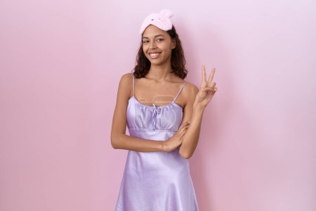 Photo for Young hispanic woman wearing sleep mask and nightgown smiling with happy face winking at the camera doing victory sign with fingers. number two. - Royalty Free Image