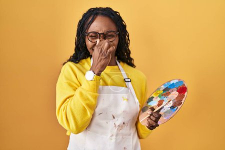 Téléchargez les photos : Femme africaine tenant palette peintre sentant quelque chose de puant et dégoûtant, odeur intolérable, retenant souffle avec les doigts sur le nez. mauvaise odeur - en image libre de droit