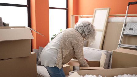 Foto de Middle age woman with grey hair unpacking cardboard box at new home - Imagen libre de derechos
