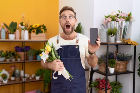 Foto de Hombre de mediana edad con barba trabajando en floristería mostrando la pantalla del teléfono inteligente celebrando loco y sorprendido por el éxito con los ojos abiertos gritando emocionado. - Imagen libre de derechos