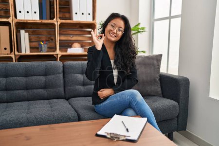 Photo for Young asian woman at consultation office smiling positive doing ok sign with hand and fingers. successful expression. - Royalty Free Image