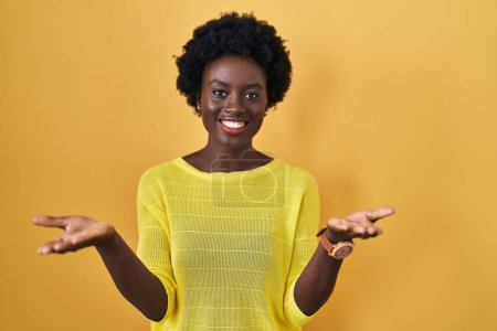 Téléchargez les photos : Jeune femme africaine debout sur jaune studio souriant joyeuse offrant des mains donnant assistance et acceptation. - en image libre de droit