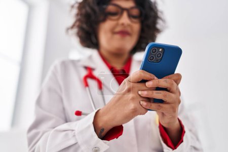 Photo for Young beautiful hispanic woman doctor using smartphone standing at clinic - Royalty Free Image