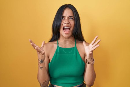 Photo for Brunette woman standing over yellow background crazy and mad shouting and yelling with aggressive expression and arms raised. frustration concept. - Royalty Free Image