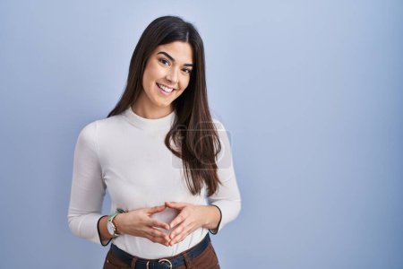 Foto de Young brunette woman standing over blue background hands together and fingers crossed smiling relaxed and cheerful. success and optimistic - Imagen libre de derechos