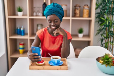 Téléchargez les photos : Young african american woman using smartphone having breakfast at home - en image libre de droit