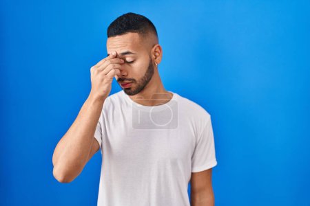 Foto de Young hispanic man standing over blue background tired rubbing nose and eyes feeling fatigue and headache. stress and frustration concept. - Imagen libre de derechos