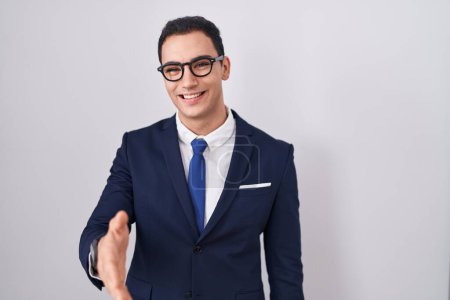 Foto de Joven hombre hispano vistiendo traje y corbata sonriente amistoso ofreciendo apretón de manos como saludo y bienvenida. negocios exitosos. - Imagen libre de derechos