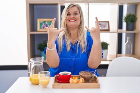 Téléchargez les photos : Caucasian plus size woman eating breakfast at home shouting with crazy expression doing rock symbol with hands up. music star. heavy concept. - en image libre de droit