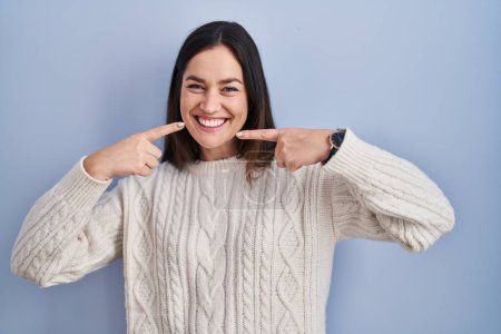 Photo for Young brunette woman standing over blue background smiling cheerful showing and pointing with fingers teeth and mouth. dental health concept. - Royalty Free Image