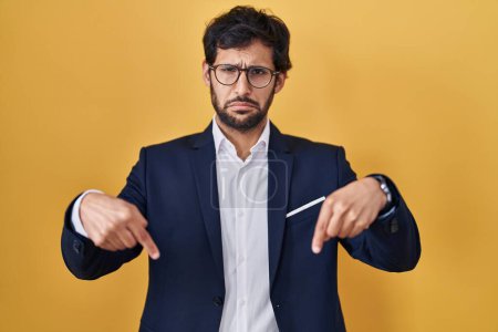 Foto de Handsome latin man standing over yellow background pointing down looking sad and upset, indicating direction with fingers, unhappy and depressed. - Imagen libre de derechos