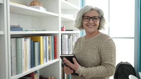 Téléchargez les photos : Femme d'âge moyen avec professeur de cheveux gris souriant livre de tenue confiant à l'université salle de classe - en image libre de droit