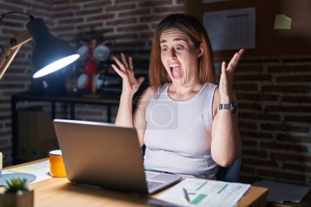 Téléchargez les photos : Femme brune travaillant au bureau la nuit célébrant fou et étonné pour le succès avec les bras levés et les yeux ouverts hurlant excités. concept gagnant - en image libre de droit