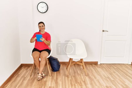 Téléchargez les photos : Jeune belle femme hispanique utilisant touchpad assis sur la chaise à la salle d'attente - en image libre de droit