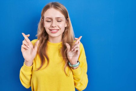 Téléchargez les photos : Young caucasian woman standing over blue background gesturing finger crossed smiling with hope and eyes closed. luck and superstitious concept. - en image libre de droit