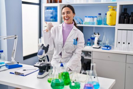 Téléchargez les photos : Jeune femme brune travaillant au laboratoire scientifique souriant avec le visage heureux regardant et pointant vers le côté avec le pouce vers le haut. - en image libre de droit