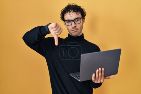 Foto de Hombre hispano trabajando con computadora portátil con cara enojada, signo negativo que muestra aversión con los pulgares hacia abajo, concepto de rechazo - Imagen libre de derechos
