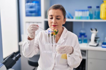 Foto de Middle age woman scientist pouring liquid on flower at laboratory - Imagen libre de derechos