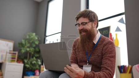 Téléchargez les photos : Young redhead man preschool teacher using laptop sitting on chair at kindergarten - en image libre de droit