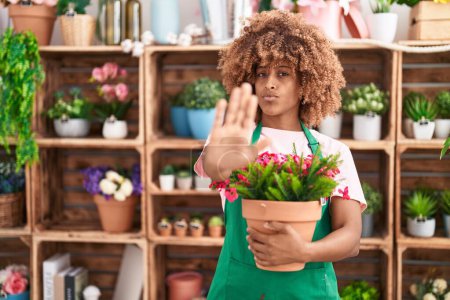 Téléchargez les photos : Jeune femme hispanique aux cheveux bouclés travaillant dans un magasin de fleuristes tenant une plante à la main ouverte faisant signe d'arrêt avec une expression sérieuse et confiante, geste de défense - en image libre de droit