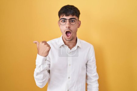 Photo for Young hispanic man standing over yellow background surprised pointing with hand finger to the side, open mouth amazed expression. - Royalty Free Image