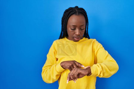 Photo for Beautiful black woman standing over blue background checking the time on wrist watch, relaxed and confident - Royalty Free Image
