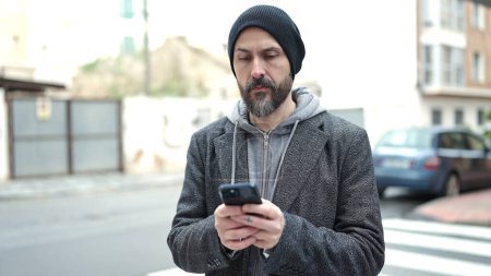 Téléchargez les photos : Jeune homme chauve utilisant un smartphone avec une expression sérieuse dans la rue - en image libre de droit