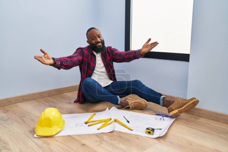Photo for African american man sitting on the floor at new home looking at blueprints looking at the camera smiling with open arms for hug. cheerful expression embracing happiness. - Royalty Free Image