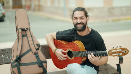 Téléchargez les photos : Young hispanic man musician playing classical guitar sitting on bench at street - en image libre de droit
