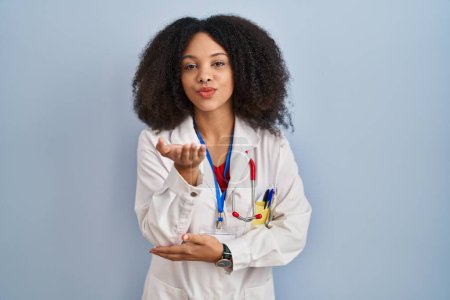Téléchargez les photos : Jeune femme afro-américaine portant l'uniforme de médecin et stéthoscope regardant la caméra souffler un baiser avec la main sur l'air étant belle et sexy. expression d'amour. - en image libre de droit