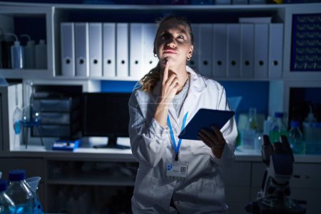 Photo for Beautiful blonde woman working at scientist laboratory late at night thinking concentrated about doubt with finger on chin and looking up wondering - Royalty Free Image