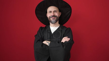 Foto de Joven calvo vestido con traje de mago de pie con los brazos cruzados gesto sobre fondo rojo aislado - Imagen libre de derechos