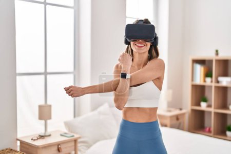 Téléchargez les photos : Jeune belle femme hispanique étirant les bras à l'aide de lunettes de réalité virtuelle dans la chambre - en image libre de droit