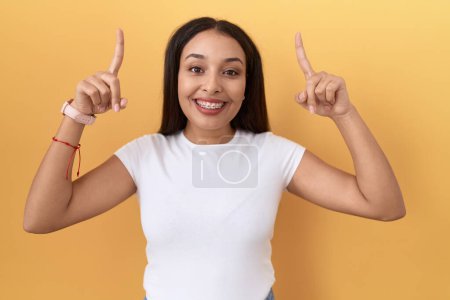 Photo for Young arab woman wearing casual white t shirt over yellow background smiling amazed and surprised and pointing up with fingers and raised arms. - Royalty Free Image