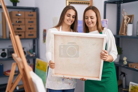 Foto de Dos mujeres artistas sonriendo confiadas sosteniendo el lienzo de dibujo en el estudio de arte - Imagen libre de derechos