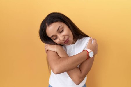 Photo for Young arab woman wearing casual white t shirt over yellow background hugging oneself happy and positive, smiling confident. self love and self care - Royalty Free Image
