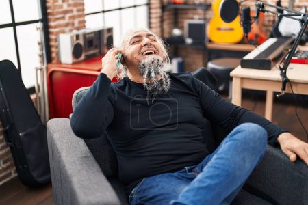 Foto de Middle age grey-haired man musician talking on smartphone sitting on chair at music studio - Imagen libre de derechos