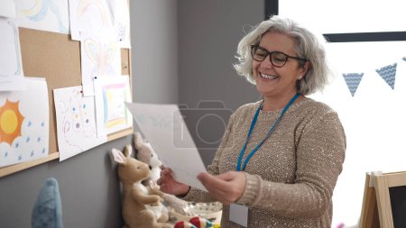 Téléchargez les photos : Middle age woman with grey hair preschool teacher smiling confident looking draw at kindergarten - en image libre de droit