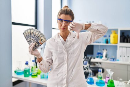 Photo for Young woman working at scientist laboratory holding money with angry face, negative sign showing dislike with thumbs down, rejection concept - Royalty Free Image