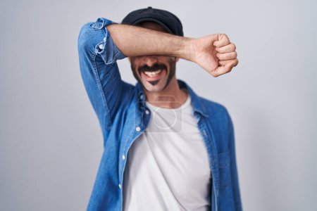 Téléchargez les photos : Homme hispanique avec barbe debout sur fond isolé couvrant les yeux avec le bras souriant gai et drôle. concept aveugle. - en image libre de droit
