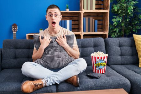 Téléchargez les photos : Young man eating popcorn afraid and shocked with surprise and amazed expression, fear and excited face. - en image libre de droit