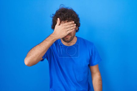 Photo for Hispanic young man standing over blue background covering eyes with hand, looking serious and sad. sightless, hiding and rejection concept - Royalty Free Image