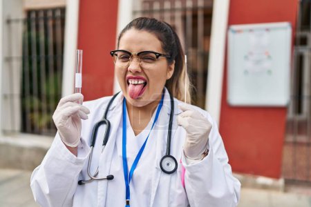 Foto de Joven doctora hispana haciendo prueba nasal de infección por coronavirus sacando la lengua feliz con expresión divertida. - Imagen libre de derechos