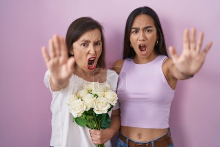 Téléchargez les photos : Mère et fille hispaniques tenant un bouquet de fleurs blanches faisant un geste d'arrêt avec les paumes des mains, expression de colère et de frustration - en image libre de droit