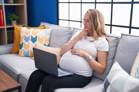 Téléchargez les photos : Jeune femme enceinte assise sur le canapé à la maison à l'aide d'un ordinateur portable visage sérieux penser à la question avec la main sur le menton, réfléchi sur l'idée déroutante - en image libre de droit