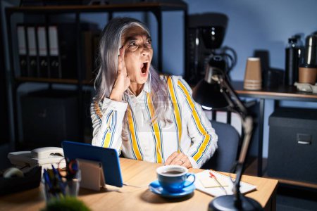 Photo for Middle age woman with grey hair working at the office at night shouting and screaming loud to side with hand on mouth. communication concept. - Royalty Free Image
