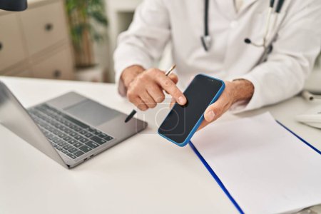 Photo for Young hispanic man wearing doctor uniform having telemedicine showing smartphone at clinic - Royalty Free Image