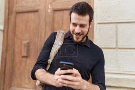 Téléchargez les photos : Jeune homme hispanique utilisant un smartphone portant un sac à dos dans la rue - en image libre de droit